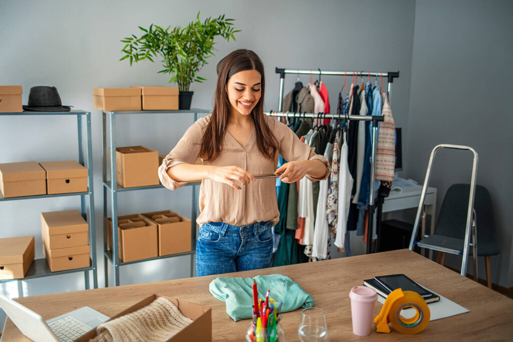 Eine junge Frau (Unternehmerin) steht an ihrem Arbeitstisch und fotografiert mit ihrem Smartphone ein Kleidungsstück. Im Hintergrund ist ein mit Schachteln und Paketen gefülltes Regal zu sehen. Der Schreibtisch ist mit diversen Büromaterialien bestückt.