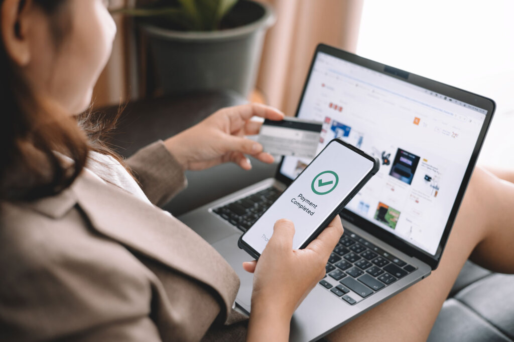 Tipps für den E-Commerce. Das Bild zeigt eine Frau, die auf einer Couch sitzt. Sie hat einen Laptop auf dem Schoß und ein Handy und eine Kreditkarte in den Händen. Auf dem Handybildschirm ist zu lesen: "Payment Completed"